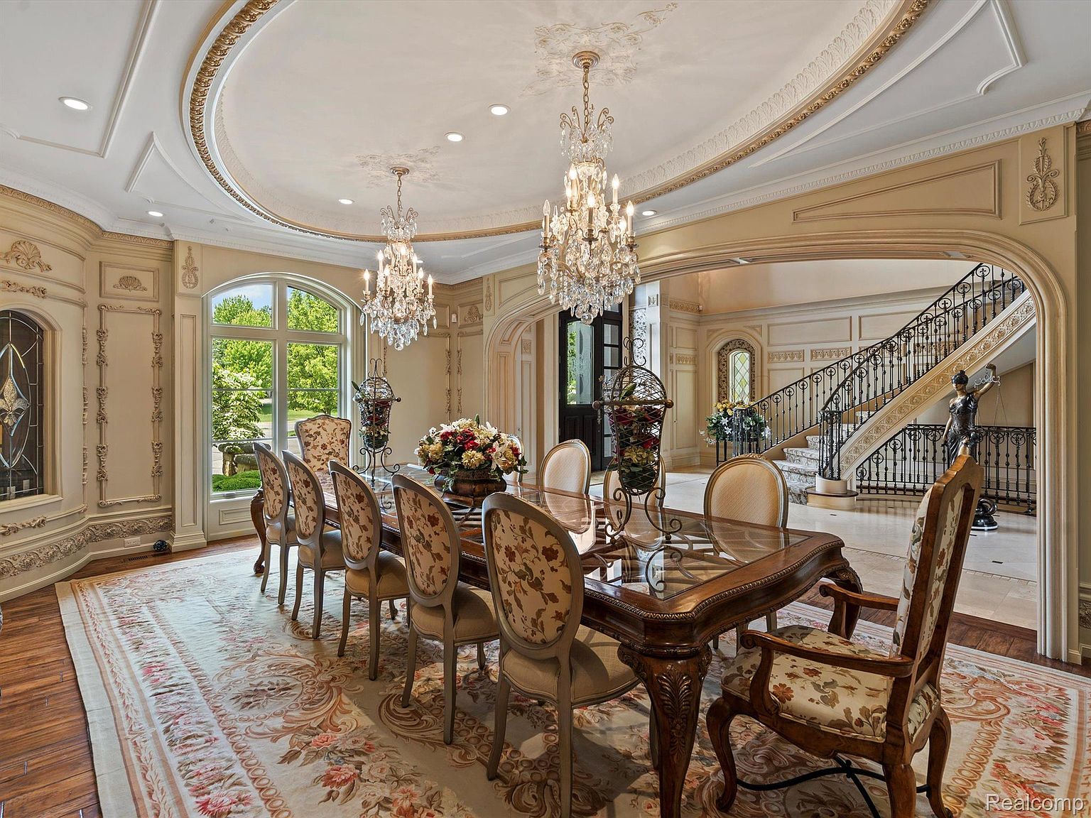 This is an opulent dining room featuring a large glass-topped dining table surrounded by ornate chairs with floral upholstery. Two crystal chandeliers hang from the decorative ceiling, and a large window provides natural light. The room opens to a grand foyer with a staircase, creating a luxurious and inviting space.