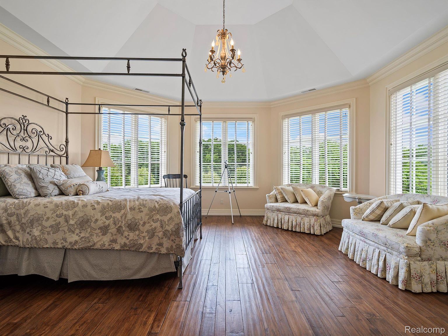 This is a primary bedroom featuring a four-poster bed with ornate metalwork and floral bedding. The room has multiple large windows with blinds, allowing natural light to illuminate the hardwood floors. Two floral sofas are placed near the windows, and a chandelier hangs from the vaulted ceiling, creating a luxurious and inviting atmosphere.