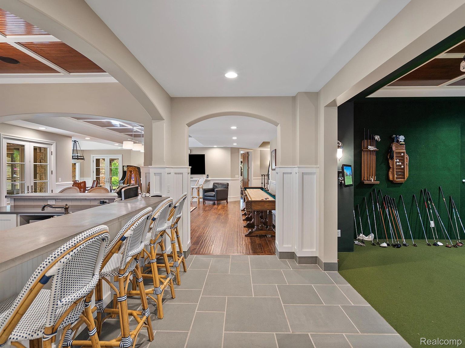 This is a view of a finished basement featuring a bar area with stylish bar stools, an open floor plan leading to a billiards area, and a golf simulator room. The flooring transitions from stone tiles to hardwood, and the walls are painted in neutral tones with white wainscoting. The space is well-lit and appears to be designed for entertainment and relaxation.