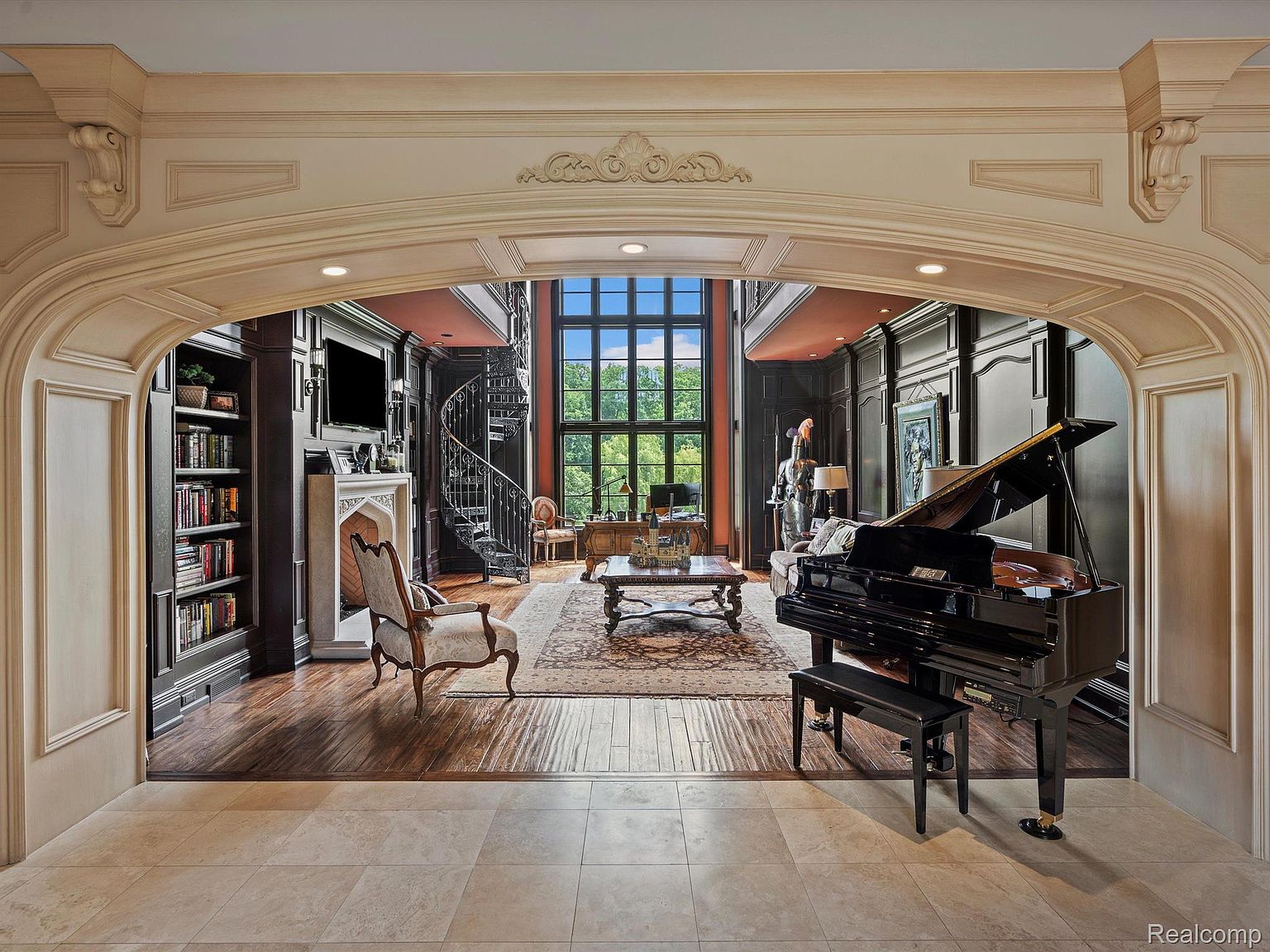 This grand living room features dark wood paneling, a spiral staircase, and a large window offering natural light. A black grand piano sits prominently in the room, complemented by an ornate rug and elegant furniture. The space exudes luxury and sophistication, perfect for entertaining or quiet relaxation.