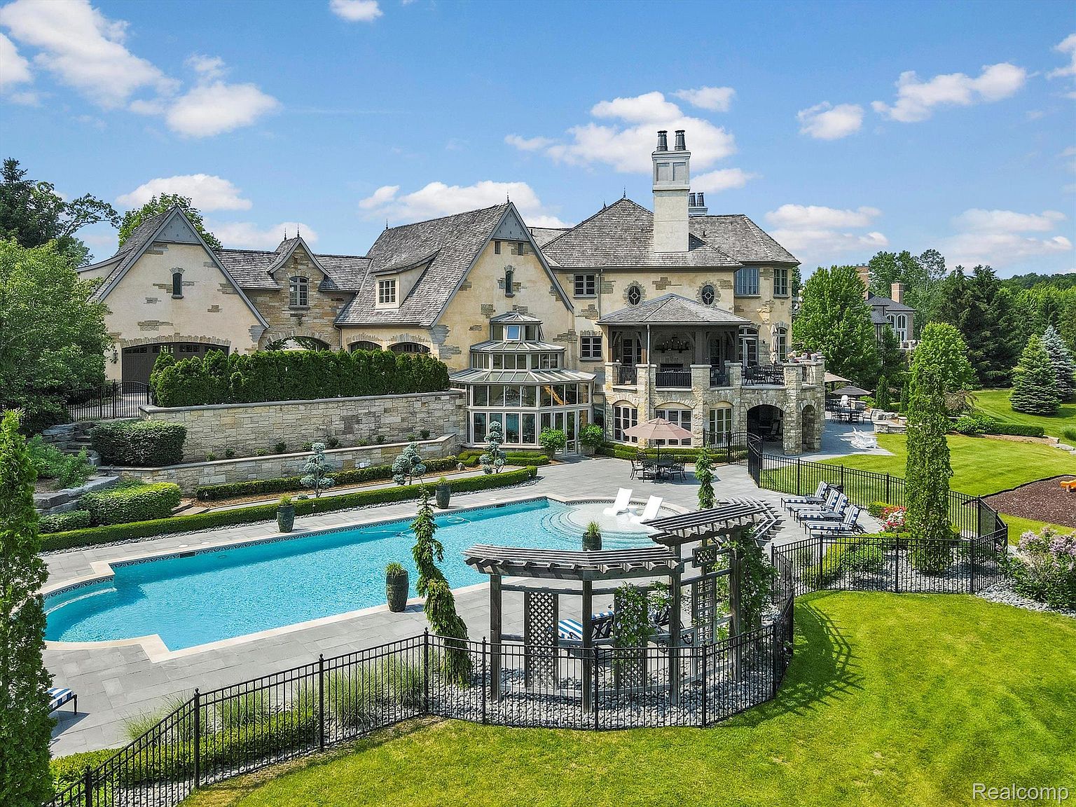 This is a stunning rear view of a luxurious estate, showcasing a large swimming pool, meticulously landscaped gardens, and an expansive patio area perfect for entertaining. The architectural style features a blend of stone and stucco, with multiple levels and outdoor living spaces, creating an inviting and opulent atmosphere. The image captures the grandeur and elegance of the property, highlighting its outdoor amenities and sophisticated design.