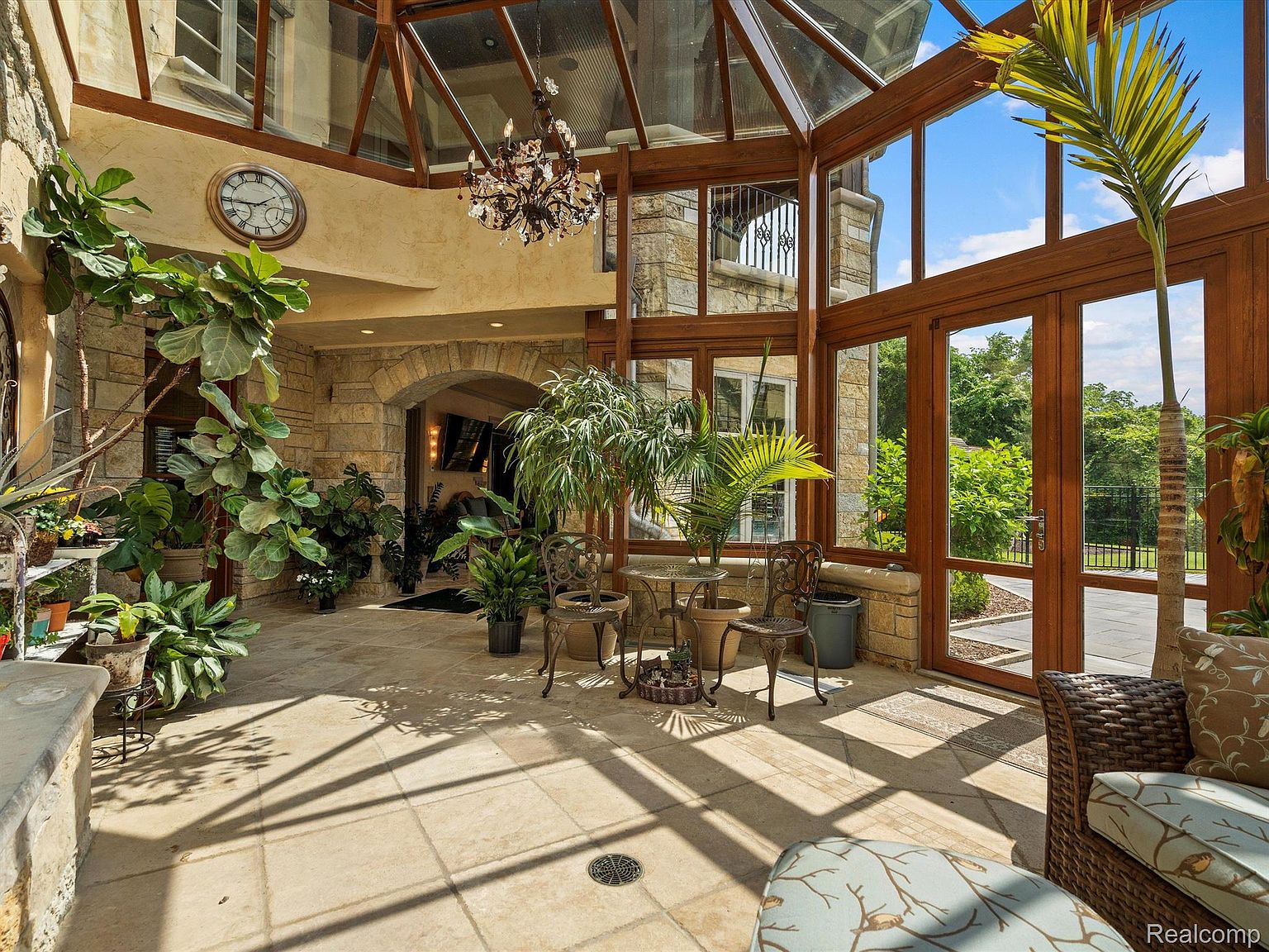 This sunroom features a glass ceiling and large windows, creating a bright and airy space filled with natural light. The room is decorated with various plants and comfortable seating, offering a relaxing indoor-outdoor feel. The stone walls and tiled floor add a touch of rustic elegance to the space.