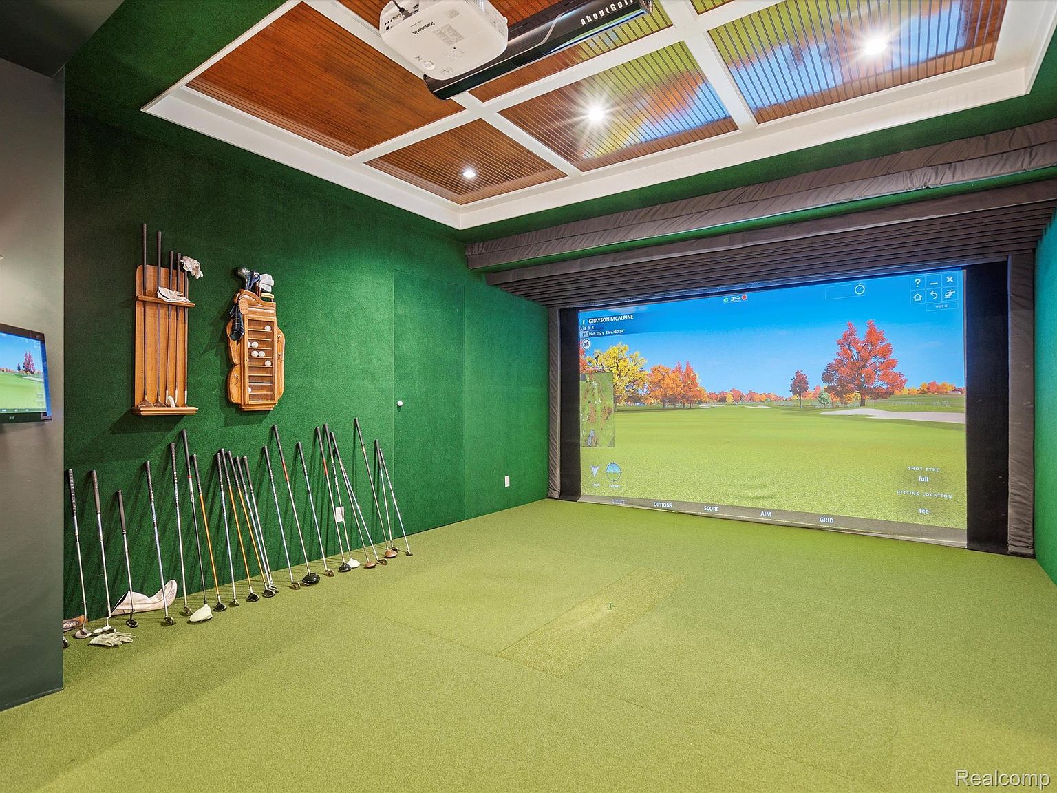 This is an indoor golf simulator room, featuring a large screen displaying a golf course, golf clubs neatly arranged along the wall, and a green carpet resembling a putting green. The room has a sophisticated design with wood paneling on the ceiling and a projector mounted above, creating an immersive golfing experience.