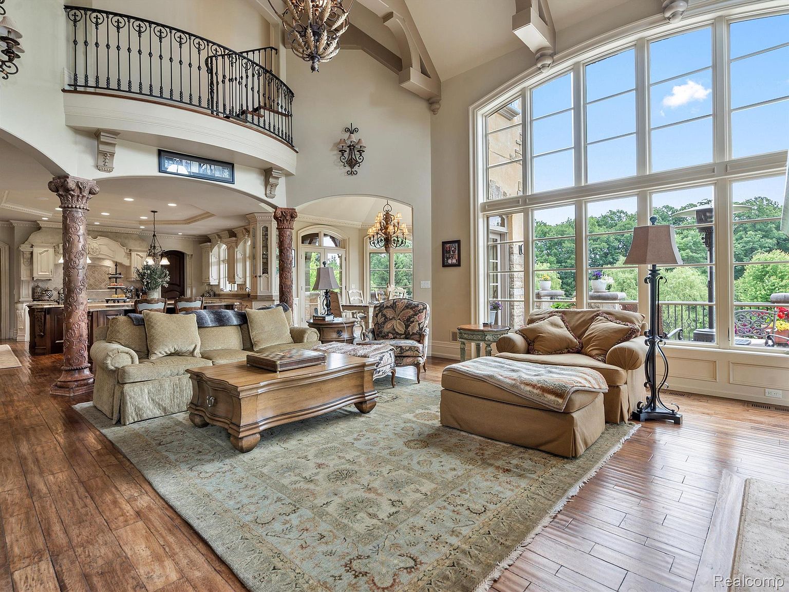 This is an interior shot of a luxurious living room featuring a soaring two-story window that floods the space with natural light. The room is furnished with comfortable, neutral-toned sofas, a large wooden coffee table, and an ornate area rug. A balcony overlooks the living area, adding to the grandeur of the space, while decorative columns and chandeliers contribute to the room's elegant and sophisticated ambiance.