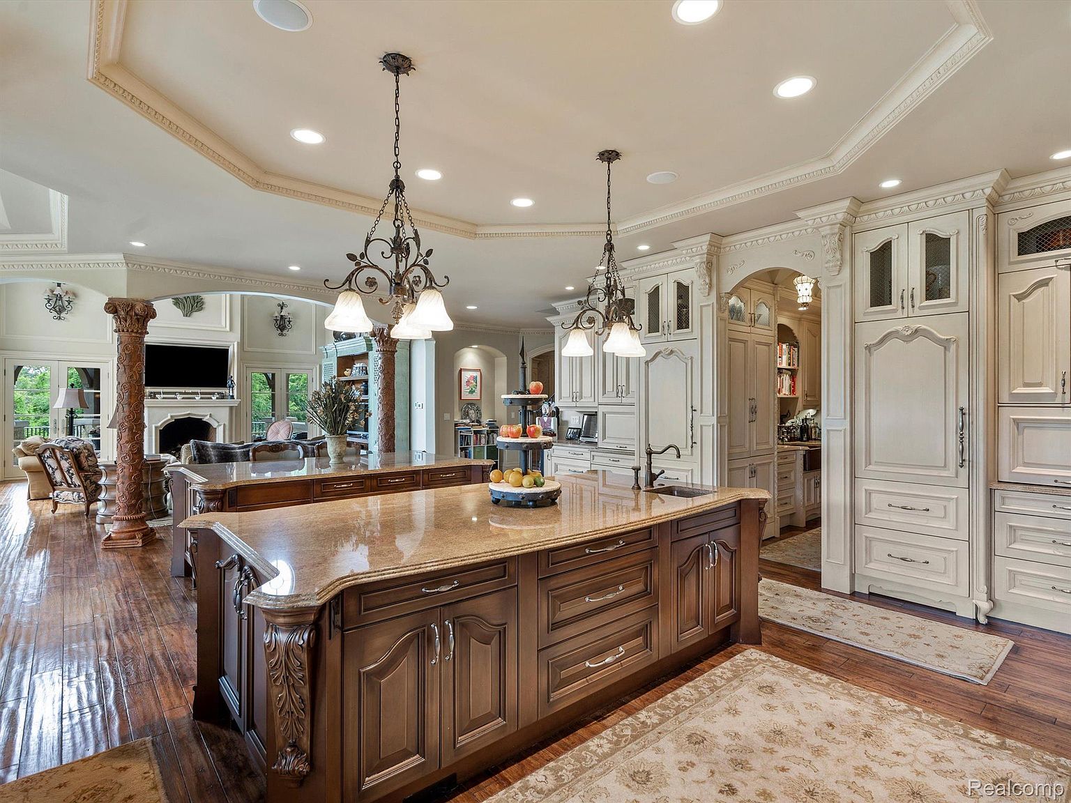 This is a spacious and luxurious kitchen featuring a large central island with a granite countertop and dark wood cabinetry. The kitchen also includes custom white cabinetry, high-end appliances, and elegant pendant lighting. The open layout connects to a living area with decorative columns, creating a seamless flow and a grand impression.