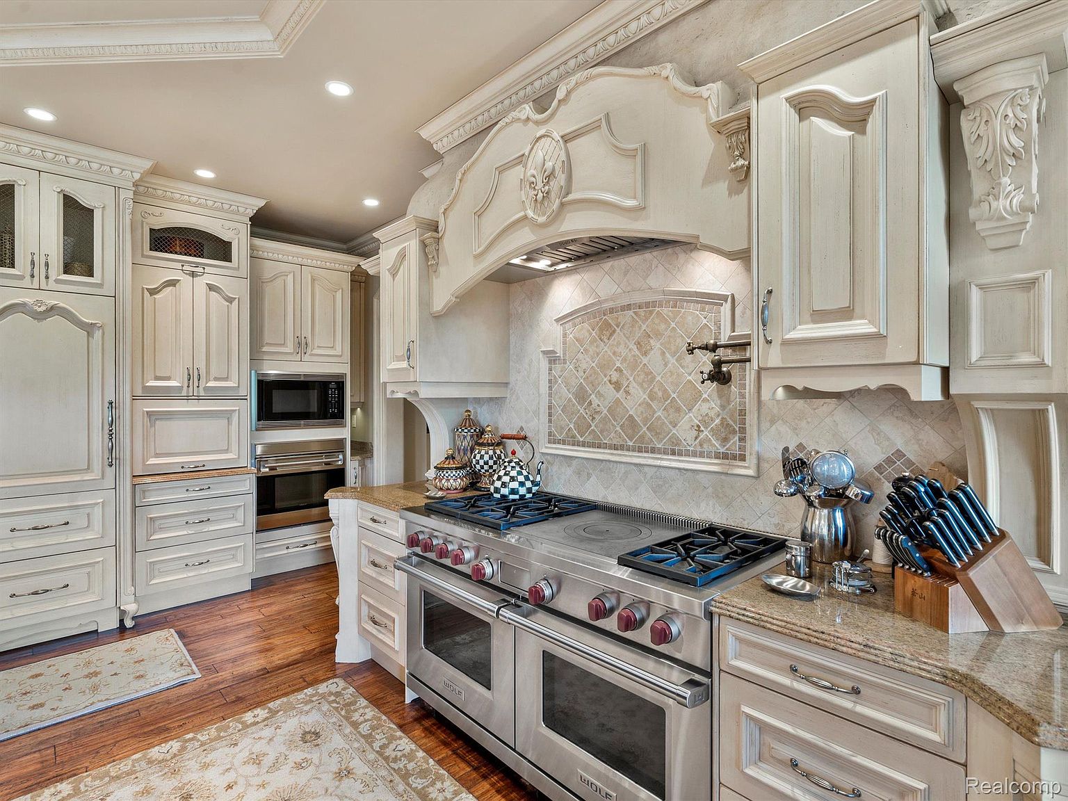 This is a well-appointed kitchen featuring custom cabinetry in an off-white finish, granite countertops, and a high-end Wolf range. The kitchen also includes a decorative range hood and tiled backsplash, adding to the luxurious feel. The overall impression is one of elegance and functionality.