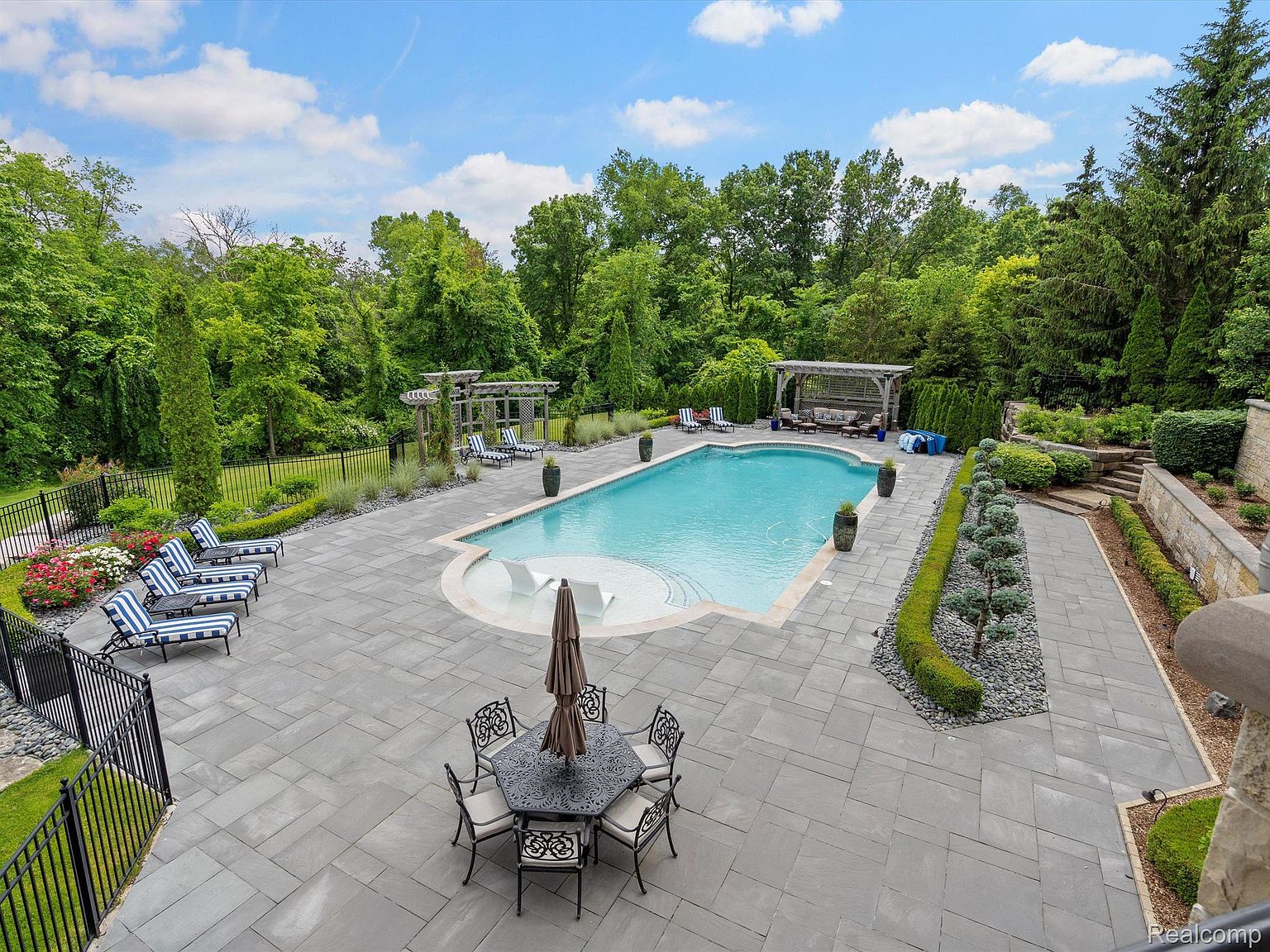 This aerial view showcases a luxurious backyard featuring a large rectangular pool with a rounded end and built-in seating. The pool is surrounded by a spacious patio made of gray pavers, complete with a dining set under an umbrella and several lounge chairs. Lush landscaping, including trees, shrubs, and decorative stone, provides privacy and enhances the overall aesthetic, creating a serene and inviting outdoor living space.