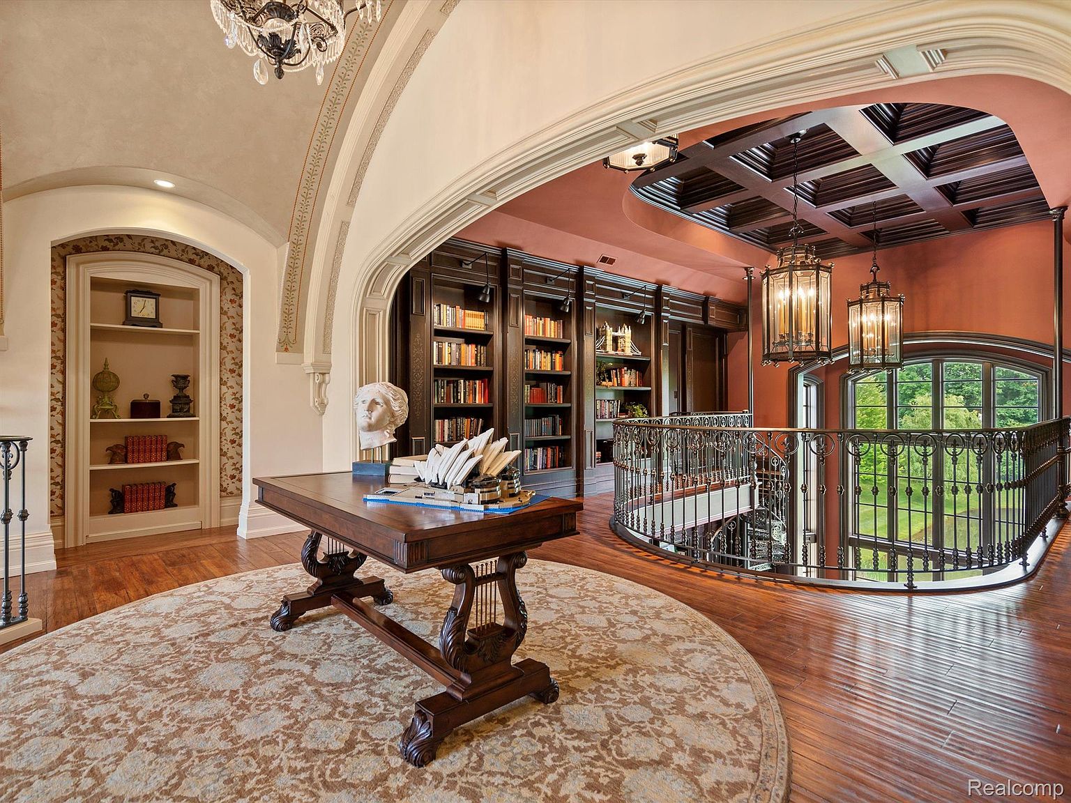 This interior shot showcases a grand hallway with a circular staircase leading to a second-floor library. The hallway features a round rug, a wooden desk with decorative elements, and arched doorways, creating a luxurious and sophisticated atmosphere. The library above has dark wood bookshelves and elegant lighting fixtures, enhancing the overall aesthetic.