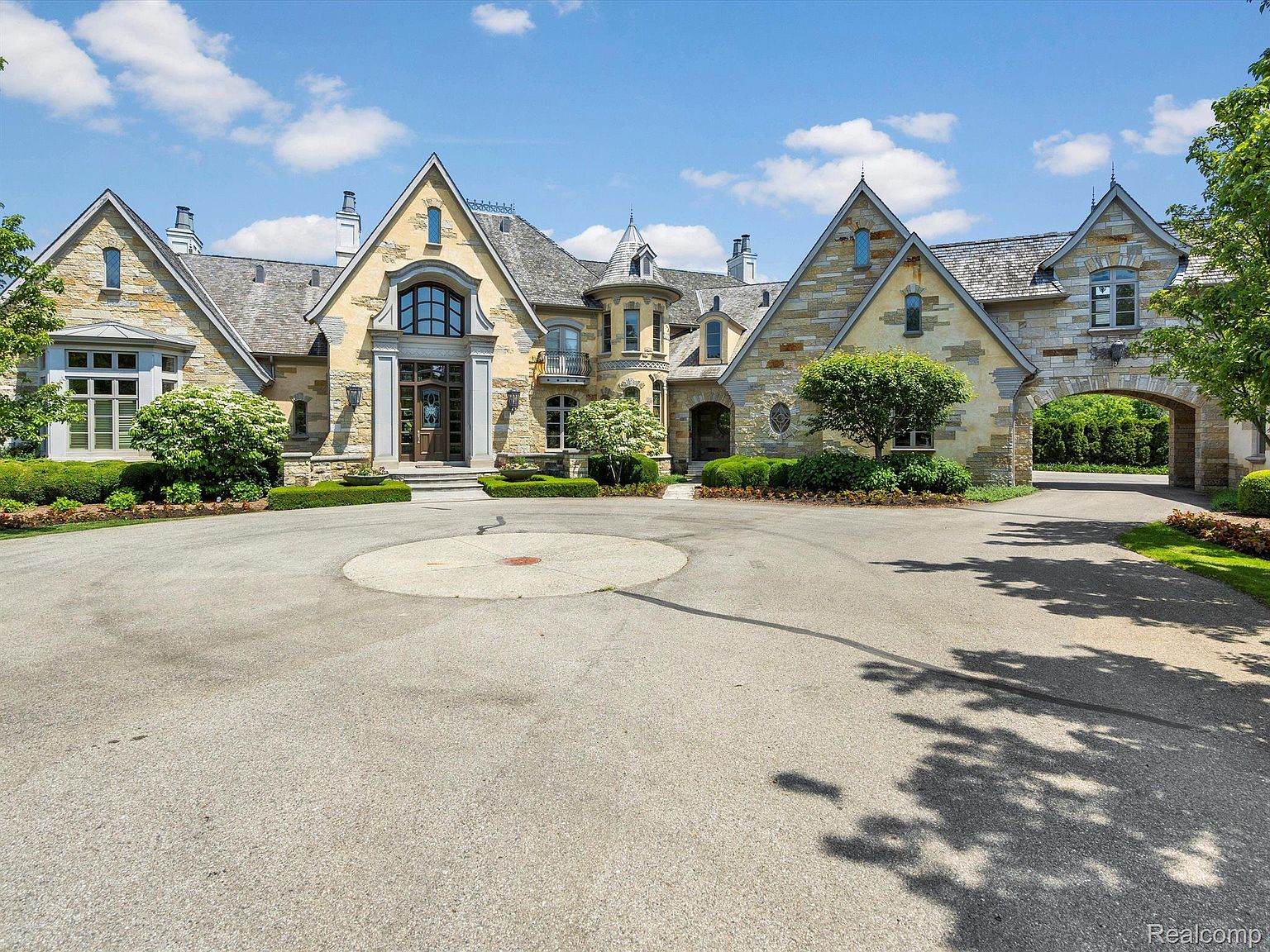 This is a front exterior view of a grand, luxurious estate. The house features a mix of stone and stucco facade, multiple gabled roofs, and an arched entryway leading to a courtyard. A circular driveway provides ample space for vehicles, and manicured landscaping enhances the property's curb appeal.