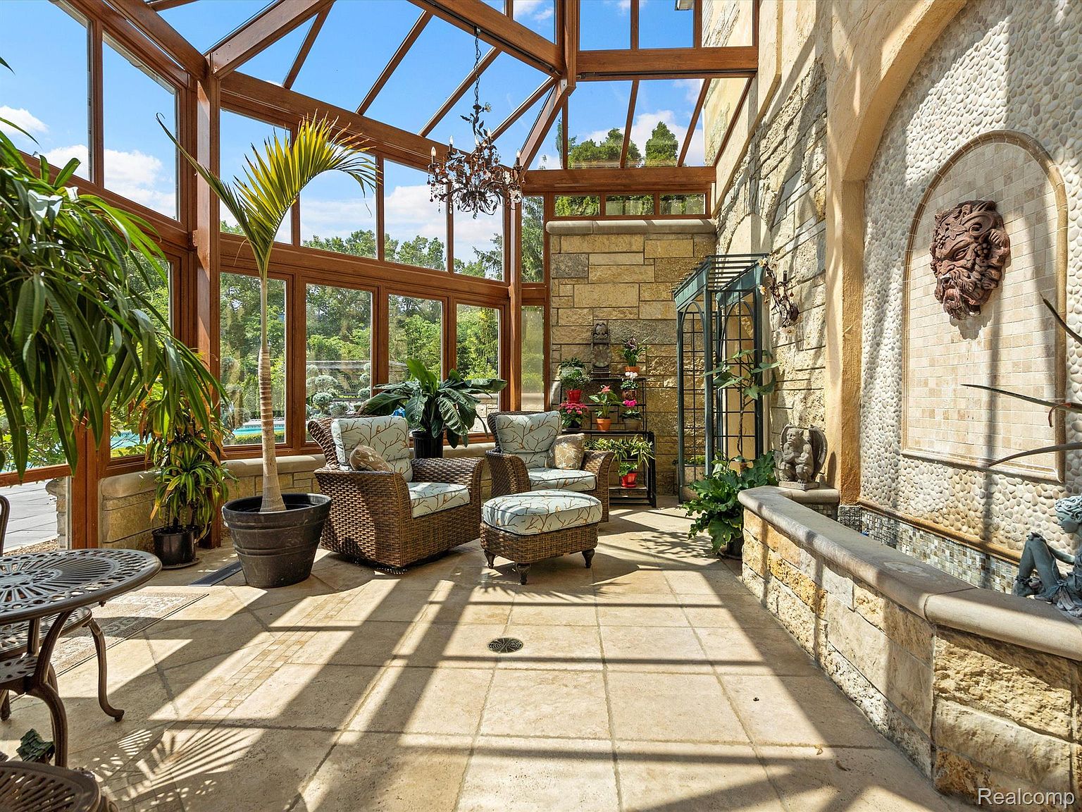 This sunroom features a glass ceiling and walls, allowing ample natural light to flood the space. Wicker furniture with patterned cushions creates a cozy seating area, complemented by potted plants and a decorative fountain. The stone walls and tiled floor add a touch of rustic elegance, making it an inviting space for relaxation.