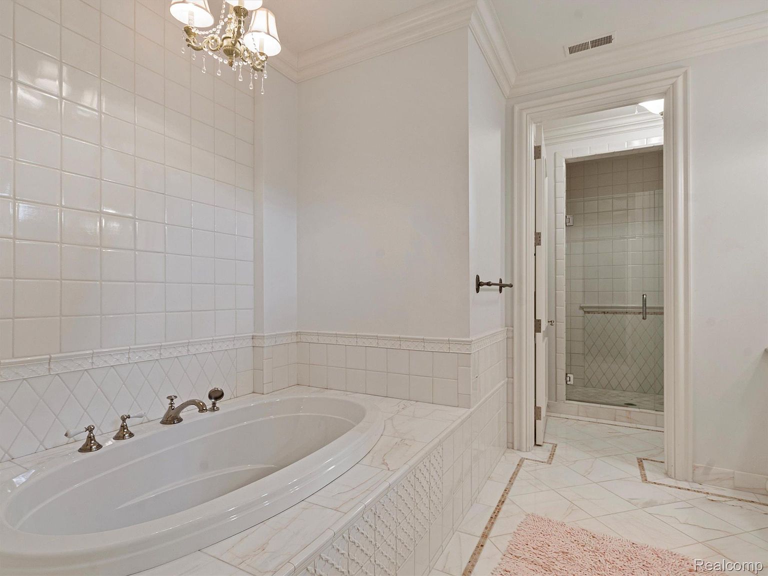 This is a primary bathroom featuring a large, oval-shaped soaking tub with elegant fixtures. The walls and tub surround are tiled in white, creating a clean and bright aesthetic. A doorway leads to a tiled shower, and the overall impression is one of luxury and relaxation.