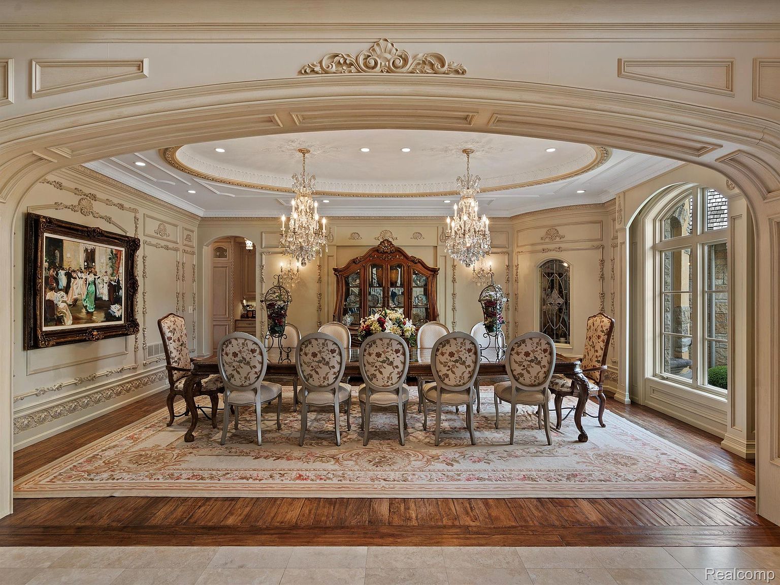 This is an opulent dining room featuring a large wooden table surrounded by ornate chairs with floral upholstery. Two crystal chandeliers hang from the decorative ceiling, illuminating the room. The walls are adorned with intricate moldings and a large painting, adding to the room's luxurious and formal atmosphere.
