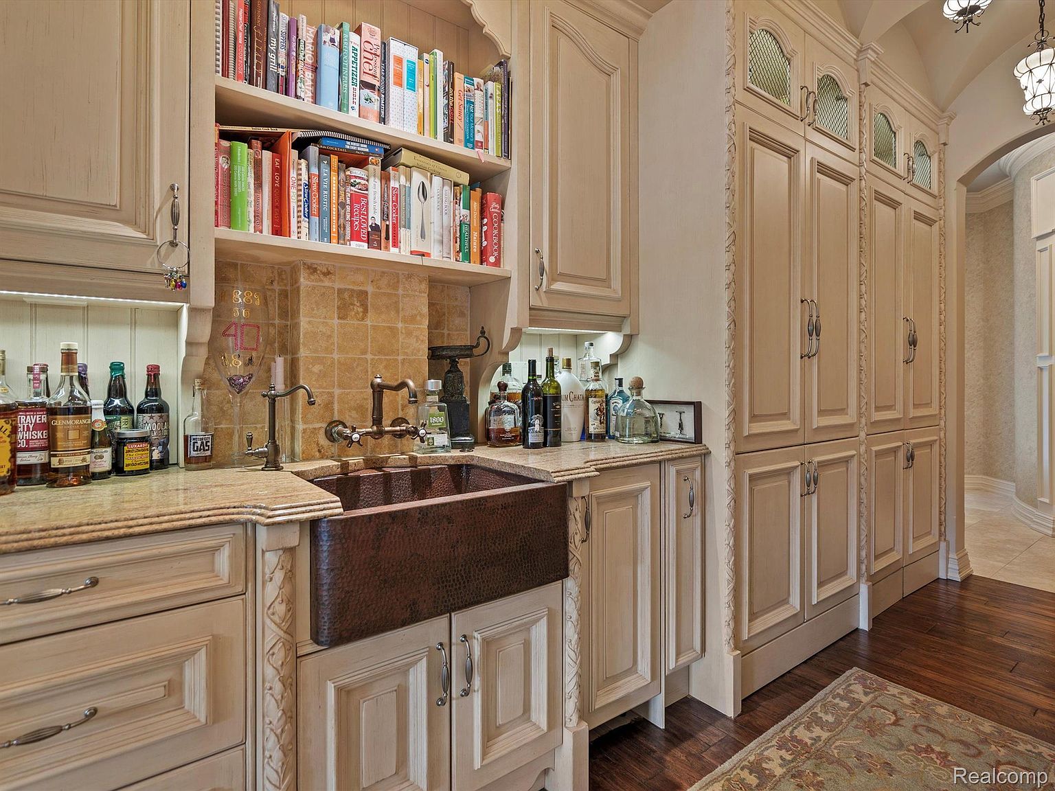This is a well-appointed kitchen featuring custom cabinetry with decorative trim, a copper farmhouse sink, and granite countertops. A built-in bookshelf above the sink adds character, while the adjacent pantry provides ample storage. The warm color palette and detailed finishes create a luxurious and inviting atmosphere.