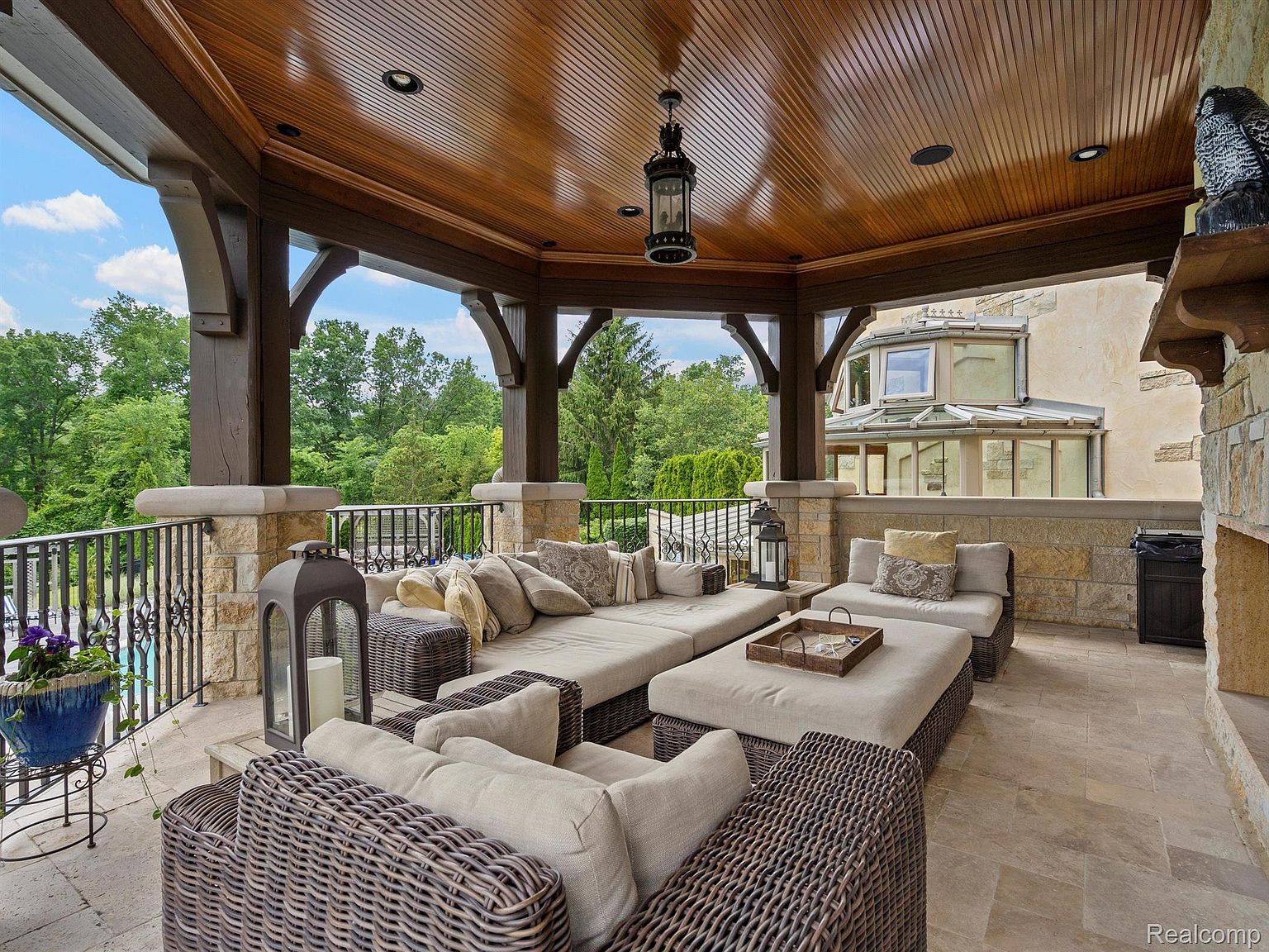 This image showcases a luxurious outdoor living space, featuring comfortable wicker seating arrangements with plush cushions, a wooden ceiling with a hanging lantern, and stone pillars. The patio offers a view of a lush green landscape and a glimpse of the adjacent building, creating an inviting and upscale atmosphere perfect for relaxation and entertaining.