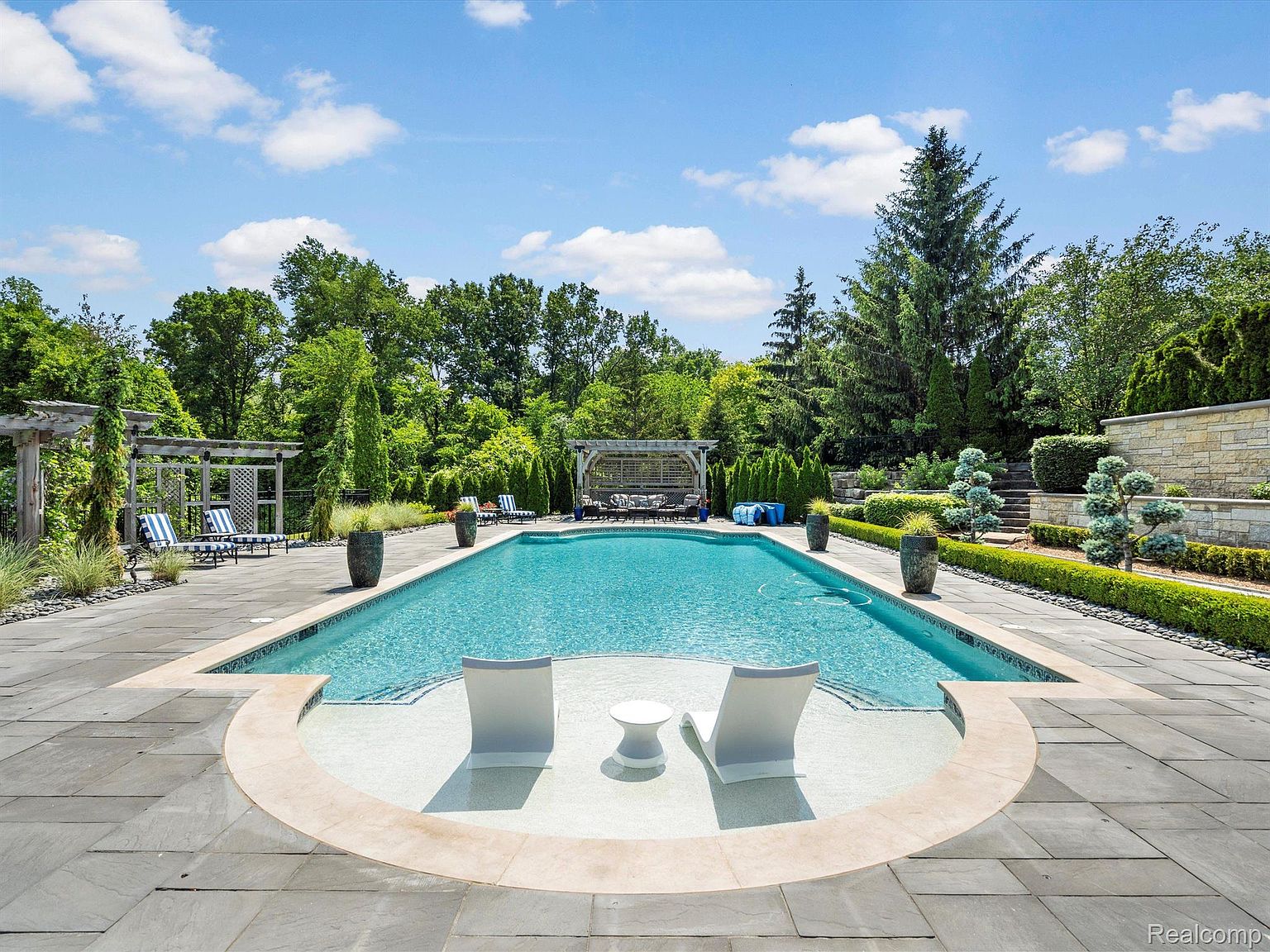 This image showcases a luxurious backyard pool area, complete with lounge chairs partially submerged in the water, a meticulously landscaped garden, and a stone patio. The pool's turquoise water contrasts beautifully with the surrounding greenery and stone elements, creating an inviting and serene atmosphere. The perspective is a wide shot, capturing the full scope of the pool area and its integration with the surrounding landscape.