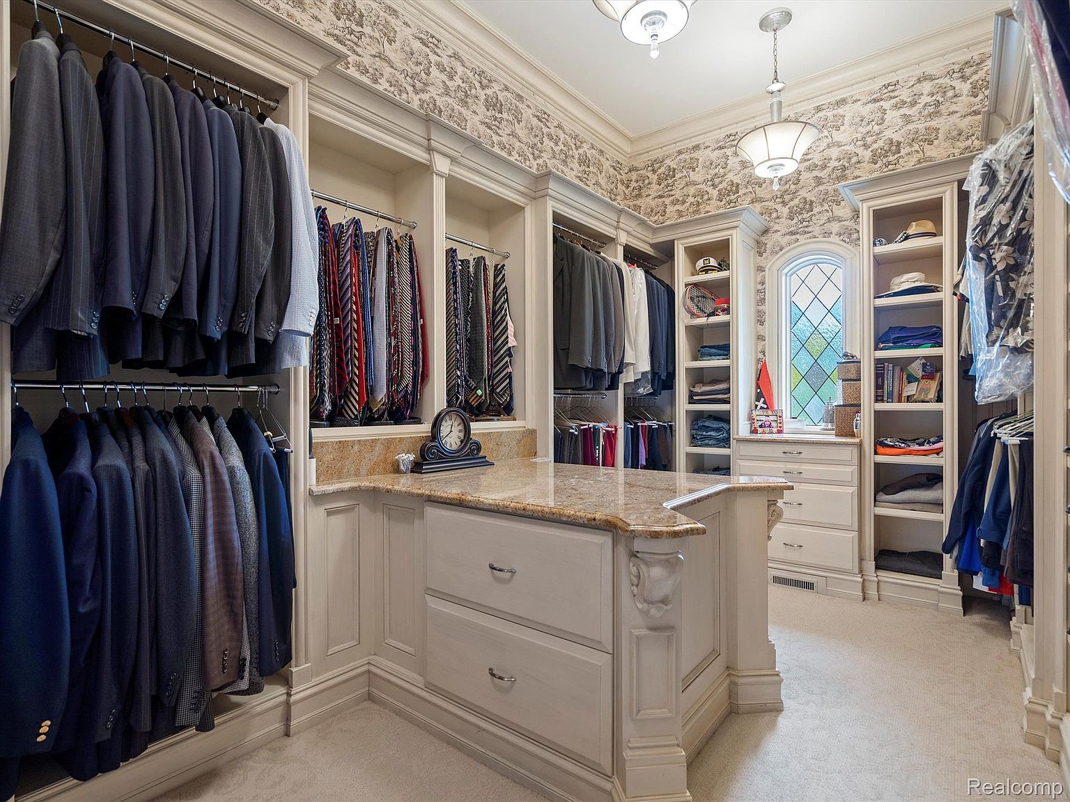 This is a spacious walk-in closet featuring custom cabinetry with ample storage for clothing and accessories. The closet includes hanging rods for suits and ties, shelving for folded items, and a central island with a granite countertop. The wallpaper adds a touch of elegance, and the overall impression is one of luxury and organization.