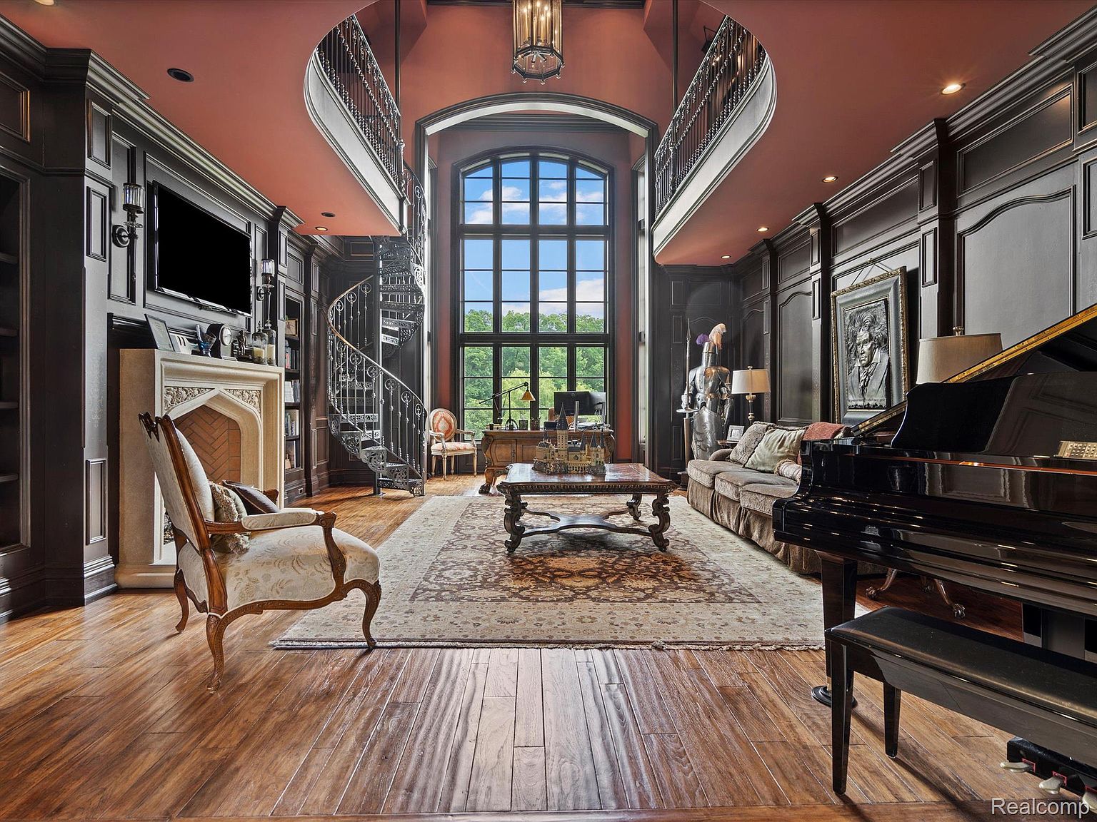 This grand living room features dark wood paneling, a soaring two-story window, and a spiral staircase leading to a balcony. A grand piano, ornate fireplace, and elegant furnishings create a luxurious and sophisticated atmosphere. The room is bathed in natural light, highlighting the intricate details of the architectural design.