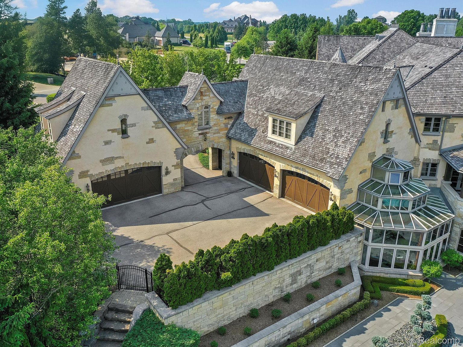 This aerial view showcases a luxurious estate with meticulously maintained grounds. The architecture features a blend of stone and stucco, complemented by a gray shingle roof. A unique glass-enclosed conservatory adds a touch of elegance, while the multiple garage bays suggest ample parking and storage.