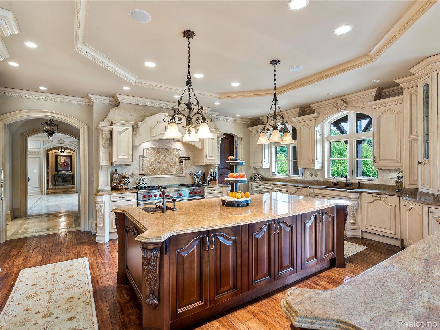 This is a luxurious kitchen featuring a large central island with dark wood cabinetry and a light-colored countertop. The kitchen also includes custom white cabinetry, granite countertops, and high-end appliances. Ornate chandeliers hang above the island, and the space is well-lit with recessed lighting, creating a warm and inviting atmosphere.