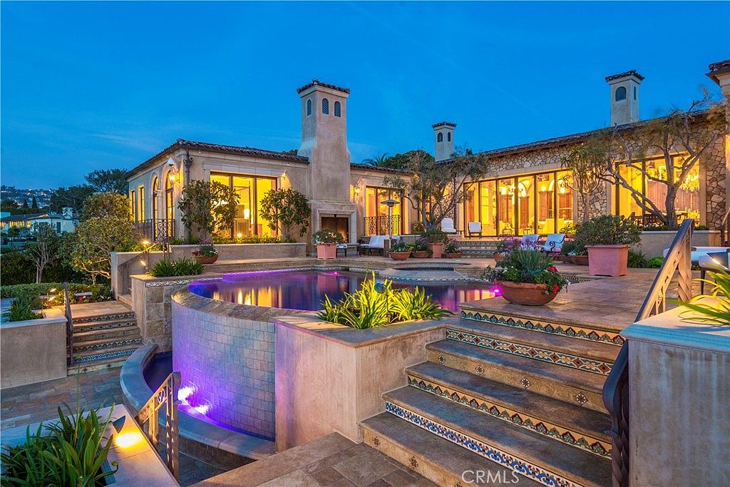 This luxurious Mediterranean-style estate features a stunning backyard oasis at dusk, highlighted by a custom-shaped pool with integrated purple accent lighting. The multi-level stone patio includes elegant stairs adorned with decorative patterned tiles, leading up to a spacious terrace with large windows and a prominent stone-clad chimney breast. The scene is framed by lush landscaping and warm exterior lighting, creating a sophisticated and inviting atmosphere for outdoor entertaining.