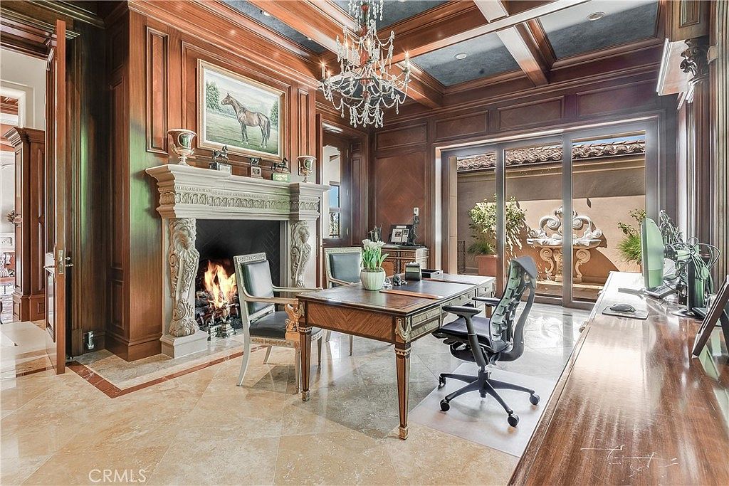 This sophisticated home office features rich wood-paneled walls, a coffered ceiling, and a grand stone fireplace with an ornate mantel. A large executive desk sits in the center of the room, complemented by a crystal chandelier and polished stone flooring. Large glass doors provide a view into a private courtyard, creating a seamless transition between the opulent interior and the outdoor space.