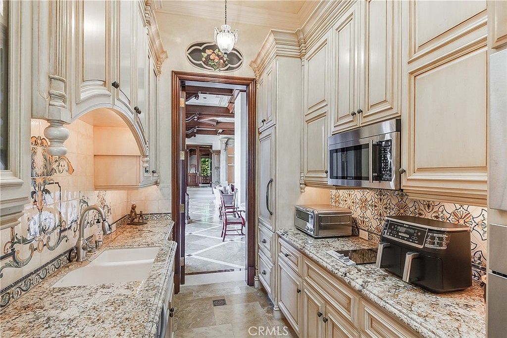 This galley-style kitchen features ornate, light-toned cabinetry with intricate detailing and stone countertops. A decorative tile backsplash adds a patterned accent, while a double-basin sink and built-in microwave are integrated into the workspace. The perspective looks through the kitchen toward an adjacent living area, highlighting the transition between spaces.