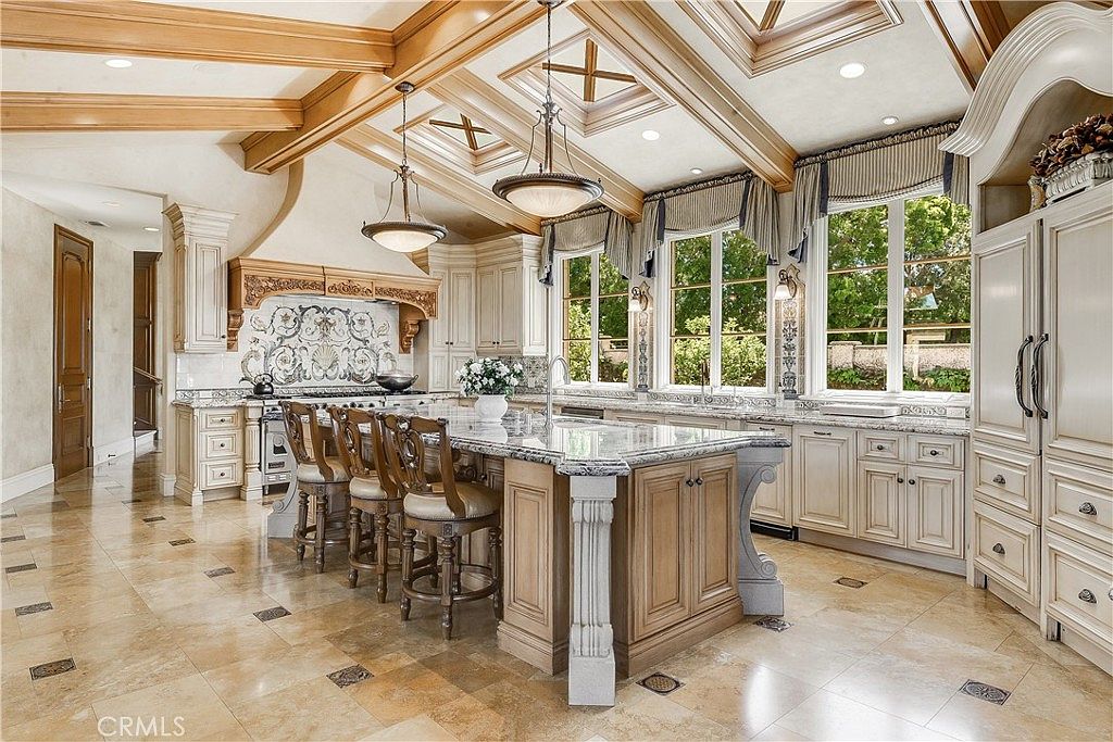This expansive, traditional-style kitchen features a large central island with bar seating and a stone countertop, serving as the focal point of the room. The space is defined by intricate wood-beamed ceilings, custom cabinetry with ornate detailing, and a decorative tile backsplash behind the range. Large windows with valances allow natural light to fill the room, highlighting the polished stone flooring and elegant pendant lighting.