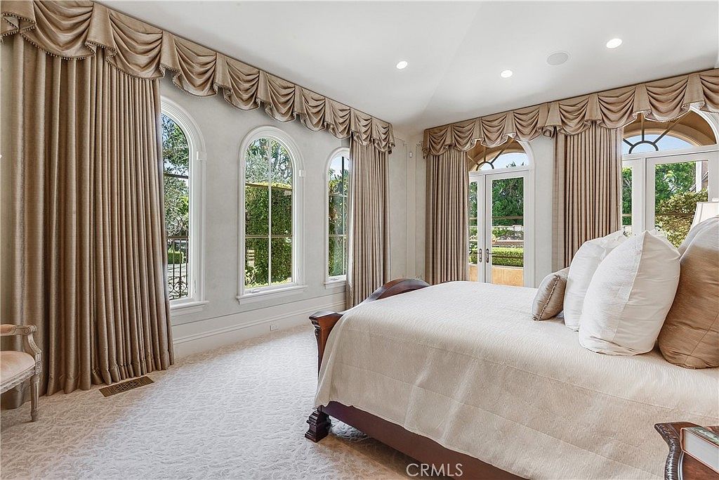 This elegant primary bedroom features a large, dark wood-framed bed topped with plush, neutral-toned bedding. The room is bathed in natural light from a series of tall, arched windows adorned with elaborate, draped valances and floor-length curtains. The space is finished with light-colored carpeting and a glass door that provides direct access to an outdoor area, creating a bright and sophisticated atmosphere.