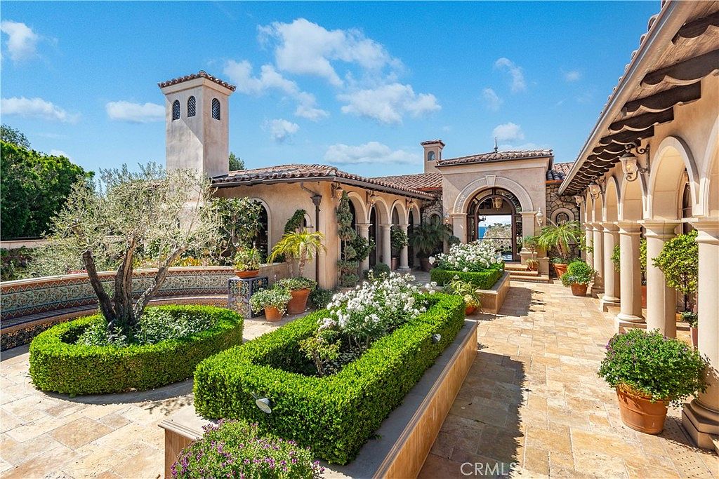 This elegant Mediterranean-style courtyard features a stone-paved patio surrounded by manicured boxwood hedges and lush landscaping. The space is defined by a series of arched colonnades and a prominent tower, creating a sophisticated outdoor living area that leads toward a scenic coastal view. The warm, sun-drenched atmosphere is enhanced by vibrant flowers and a mature olive tree, offering a serene and luxurious retreat.