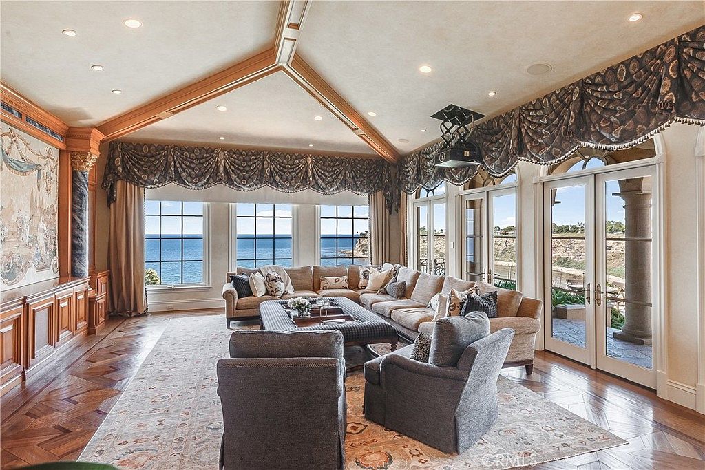 This expansive living room features a vaulted ceiling with exposed wood beams and a large, neutral-toned sectional sofa arranged around a central ottoman. The space is defined by its panoramic ocean views framed by large windows and glass doors that lead to an outdoor area. A patterned area rug sits atop rich, dark wood flooring, while decorative valances add a traditional touch to the windows.