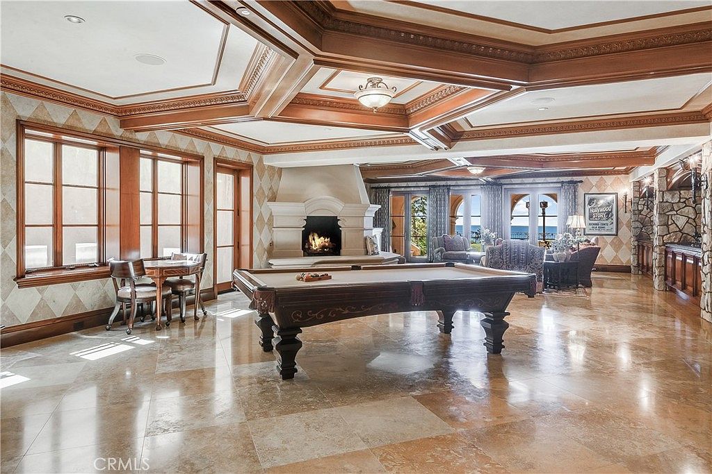 This expansive game room features a prominent billiards table as its centerpiece, set against a backdrop of polished stone flooring and intricate coffered wood ceilings. A large, ornate stone fireplace serves as a focal point, while a cozy seating area near the windows offers views of the outdoors. The room is decorated with diamond-patterned wall accents and warm wood trim, creating a sophisticated and inviting atmosphere for entertainment.
