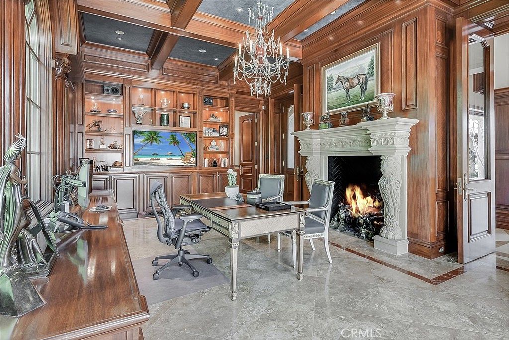 This sophisticated home office features extensive wood-paneled walls, built-in shelving, and a coffered ceiling with a central crystal chandelier. A large stone fireplace with an ornate mantel serves as a focal point, while a central desk and leather chairs provide a functional workspace. The room is finished with polished stone flooring and large windows that allow natural light to fill the space.