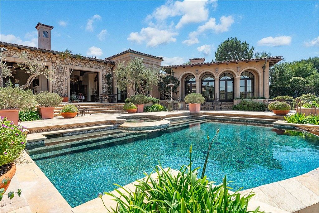 This luxurious Mediterranean-style estate features a sprawling backyard centered around a custom-shaped swimming pool with integrated spa steps. The exterior showcases a mix of textured stone masonry and elegant arched doorways, complemented by manicured landscaping and mature olive trees. The perspective captures the serene outdoor living space from the pool deck, highlighting the seamless transition between the home's architecture and the resort-like water feature.