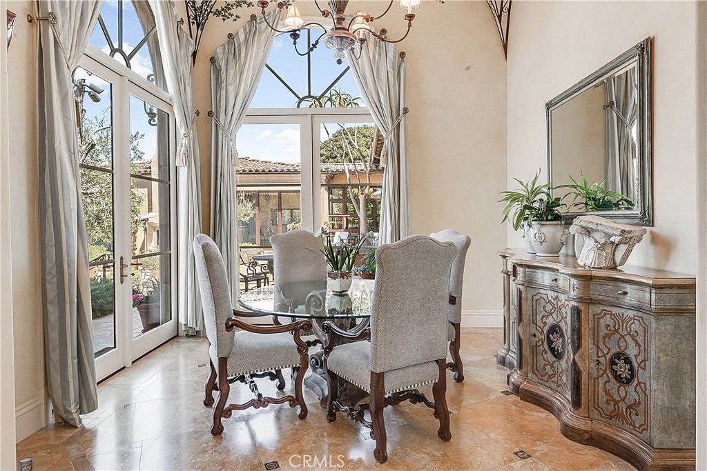 This elegant dining room features a glass-topped table surrounded by four upholstered chairs, set against a backdrop of tall windows with draped curtains. To the right, an ornate, carved wooden sideboard topped with a mirror and decorative plants adds a touch of traditional sophistication. The space is finished with light-toned stone flooring and a classic chandelier, creating a bright and refined atmosphere.