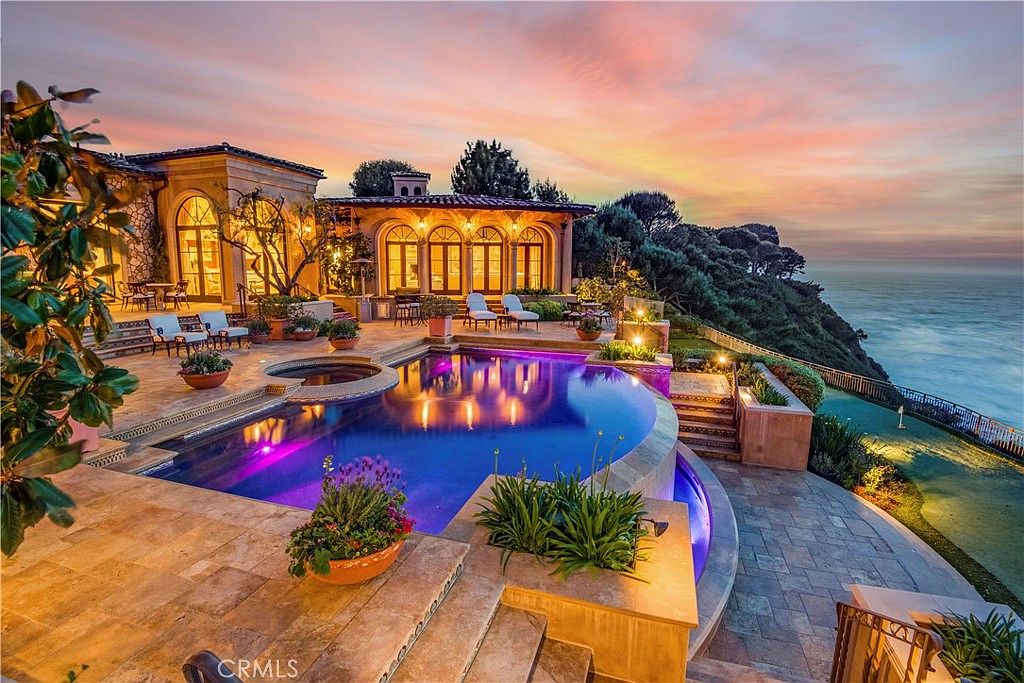 This stunning luxury estate features a multi-level stone patio centered around a curved infinity-edge pool and integrated spa, overlooking a dramatic ocean cliffside at sunset. The Mediterranean-inspired architecture includes arched glass doors and warm, ambient lighting that creates an inviting atmosphere. The expansive outdoor living space is complemented by lush landscaping and a panoramic view of the sea, offering a sophisticated and serene coastal retreat.