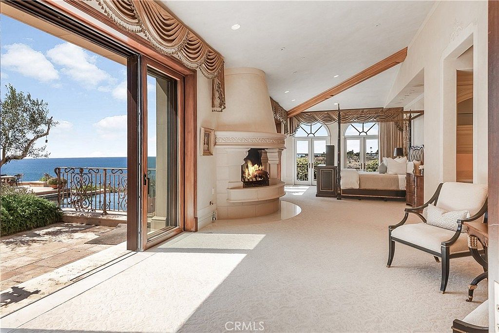 This expansive primary bedroom features a neutral color palette and a prominent, tall, light-colored stone fireplace that serves as a central focal point. The room opens directly onto a stone-paved balcony through large sliding glass doors, offering an unobstructed view of the ocean. A four-poster bed is positioned near the windows, and the space is accented by elegant drapery and a comfortable armchair, creating a luxurious and serene atmosphere.