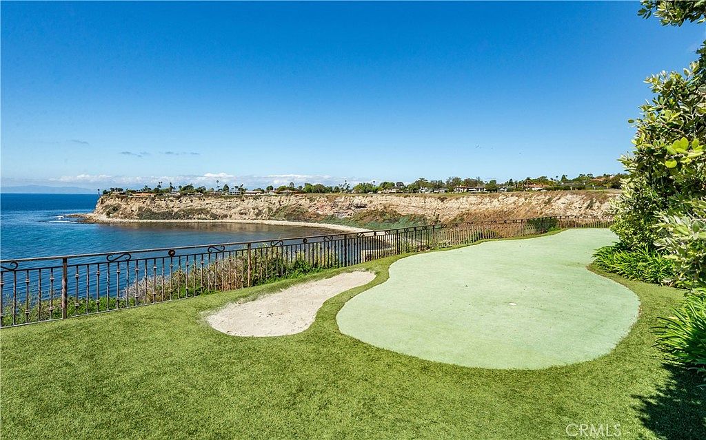This backyard features a private putting green complete with a sand bunker, set against a dramatic coastal backdrop. A decorative metal fence separates the manicured lawn from the steep cliffside overlooking the ocean and a distant bay. The scene captures a luxurious outdoor lifestyle with expansive blue skies and panoramic water views.