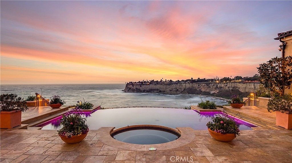 This stunning outdoor space features an infinity-edge pool and a circular spa overlooking a dramatic coastal cliffside at sunset. The patio is paved with light-toned stone tiles and accented by potted plants, creating a luxurious and serene atmosphere. The expansive ocean view and vibrant sky provide a cinematic backdrop for this high-end residential property.