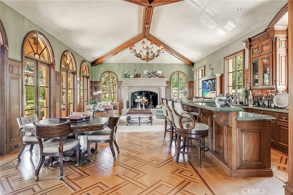 This expansive, high-ceilinged living and entertaining space features intricate patterned wood flooring and a prominent stone fireplace as a central focal point. The room includes a formal dining area on the left and a custom wood-paneled bar area on the right, all illuminated by a decorative chandelier and natural light from multiple arched glass doors. The overall aesthetic is traditional and luxurious, with rich wood cabinetry and detailed architectural molding throughout.