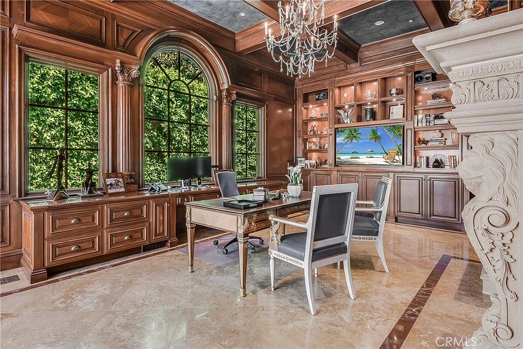 This sophisticated home office features rich, dark wood paneling throughout the walls and built-in cabinetry, creating a traditional and executive atmosphere. A central desk is positioned in the middle of the room, complemented by a crystal chandelier overhead and large arched windows that allow for ample natural light. The space is finished with polished stone flooring and a prominent stone-clad fireplace surround on the right, offering a luxurious and professional aesthetic.