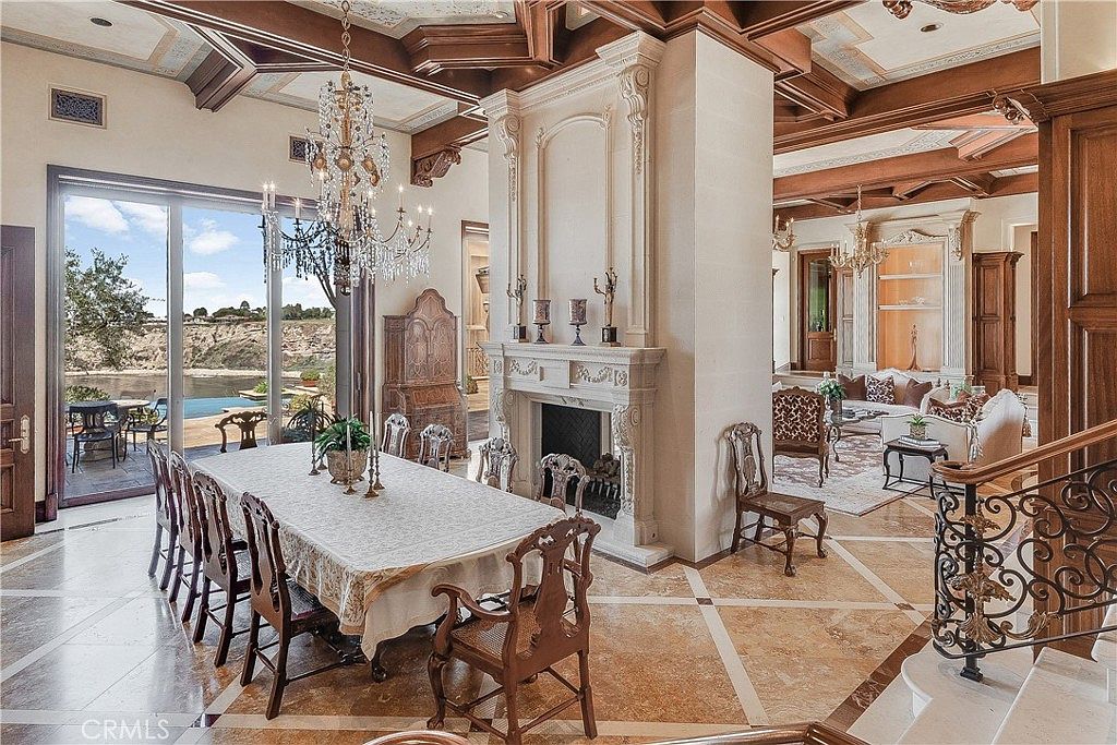 This elegant dining room features a large table set for formal gatherings, positioned beneath a grand crystal chandelier. A tall, ornate stone fireplace surround serves as a central architectural focal point, while large glass sliding doors provide a seamless transition to the outdoor patio and pool area. The space is defined by its intricate coffered wood ceiling, patterned stone flooring, and an open-concept layout that flows into an adjacent living area.