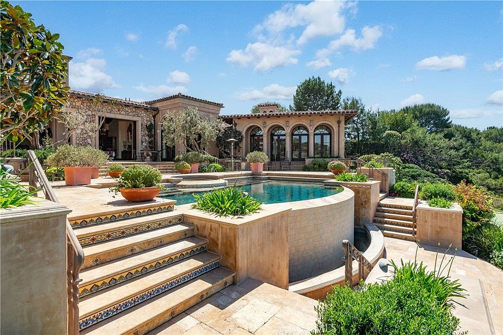 This luxurious Mediterranean-style estate features a tiered outdoor living space centered around a curved infinity-edge pool. The scene includes stone-paved terraces, decorative tile-accented staircases, and lush landscaping with potted plants, all set against a bright blue sky. The perspective captures the seamless integration of the home's architecture with the outdoor recreational area, emphasizing a high-end, resort-like atmosphere.
