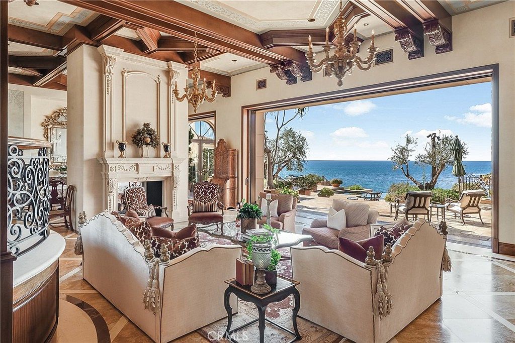 This elegant living room features a sophisticated, open-concept design with a coffered wood-beam ceiling and intricate patterned stone flooring. A grand fireplace with a detailed stone mantel serves as a focal point, while expansive glass doors slide open to reveal a seamless transition to a patio overlooking the ocean. The space is furnished with plush, neutral-toned seating arranged to maximize the panoramic coastal views.