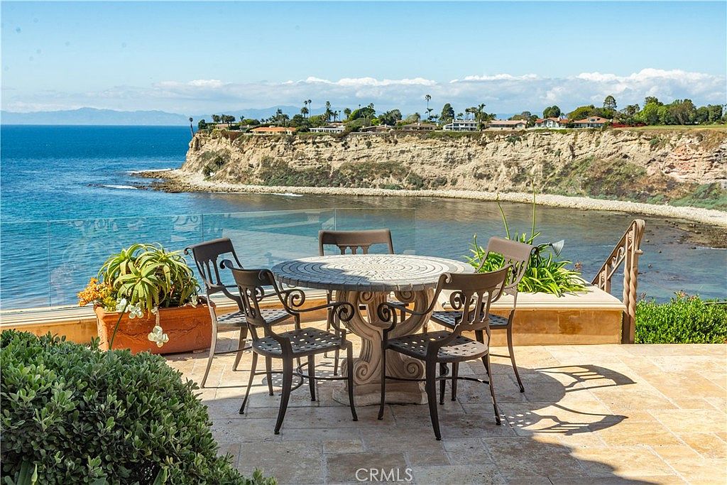 This outdoor patio space features a stone-topped dining table surrounded by four metal chairs, set against a stunning panoramic backdrop of the ocean and a rugged coastal cliff. The area is paved with light-toned stone tiles and includes a potted plant, all enclosed by a clear glass railing that preserves the expansive water view. The scene captures a serene, high-end coastal living environment perfect for al fresco dining.