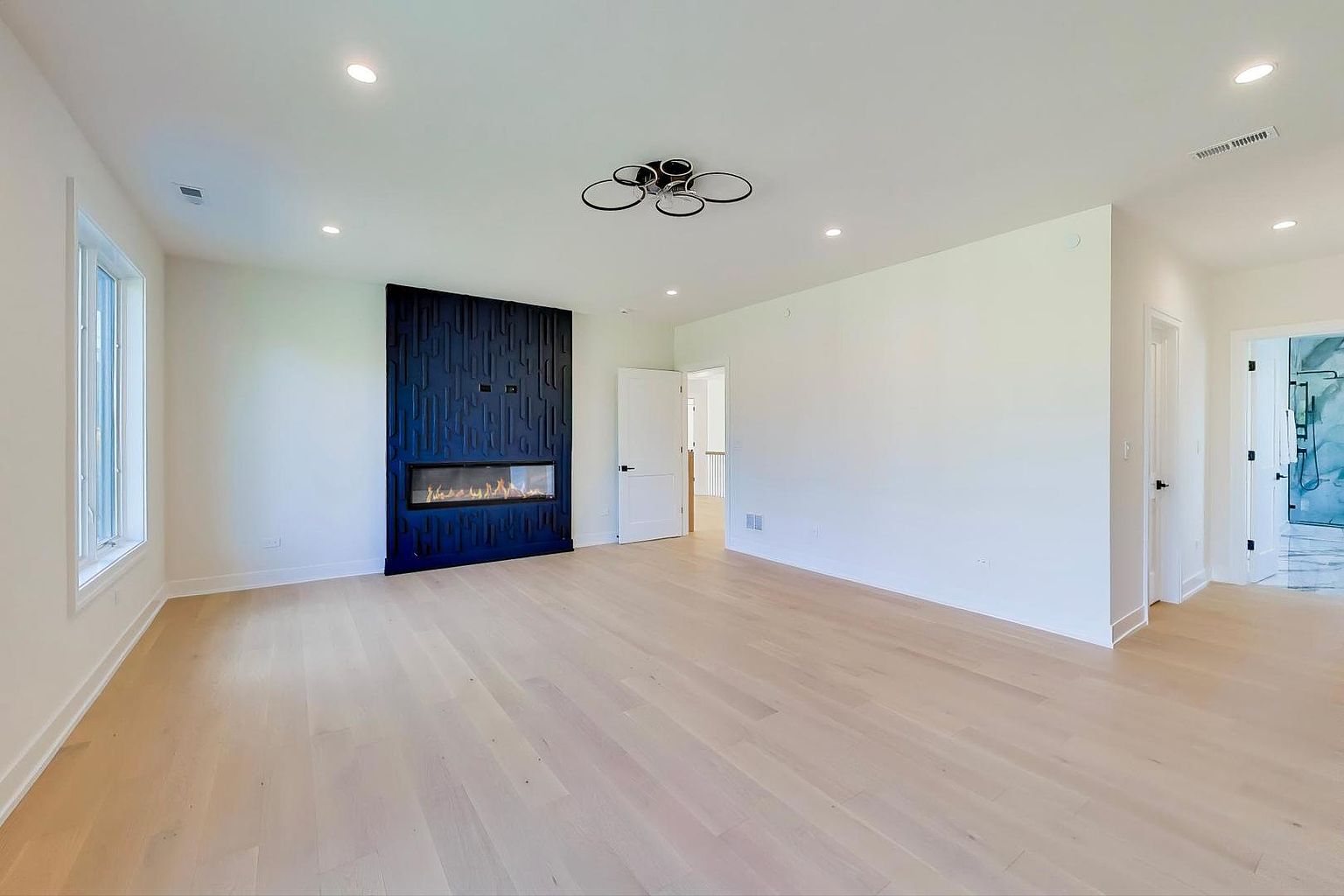 This is a spacious living room featuring light hardwood floors and white walls, creating a bright and airy atmosphere. A modern fireplace with a dark blue textured surround serves as a focal point. The room is well-lit with recessed lighting and a unique ceiling fixture, offering a clean and contemporary aesthetic.