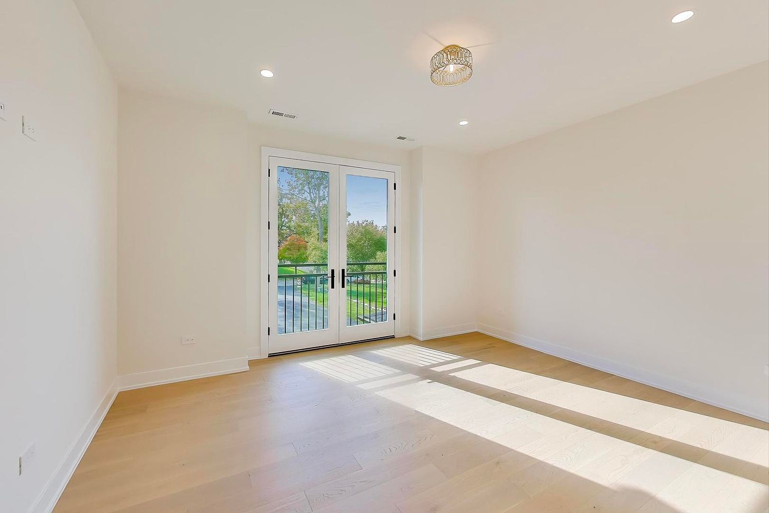 This is a bright and airy bedroom featuring light hardwood floors and white walls, creating a spacious feel. A set of French doors leads to a balcony, providing natural light and a view of the outdoors. The room is minimalist in style, emphasizing clean lines and a serene atmosphere.