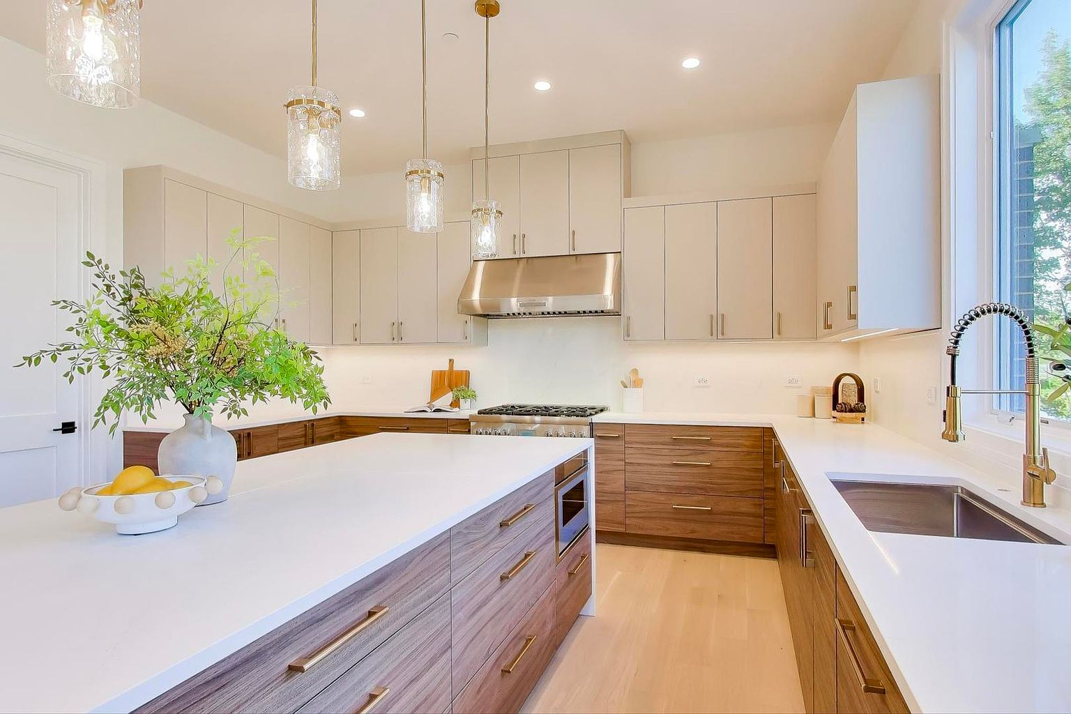 This is a bright and modern kitchen featuring a large island with a white countertop and wood-grain cabinetry. The kitchen includes stainless steel appliances, pendant lighting, and a window that provides natural light. The color palette is neutral with a mix of white and wood tones, creating a clean and inviting space.