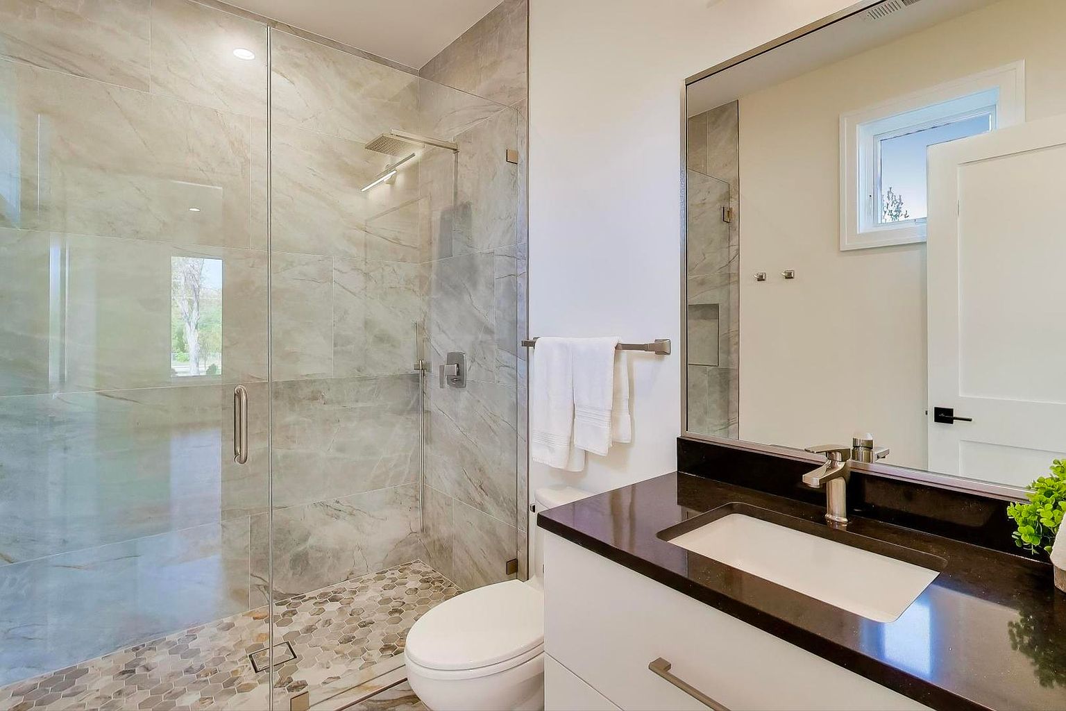This is a well-lit primary bathroom featuring a glass-enclosed shower with marble-like tile and a modern showerhead. The vanity has a dark countertop, a white sink, and white cabinets with sleek hardware. A large mirror reflects the space, and the overall design is clean and contemporary, creating a luxurious and spa-like atmosphere.