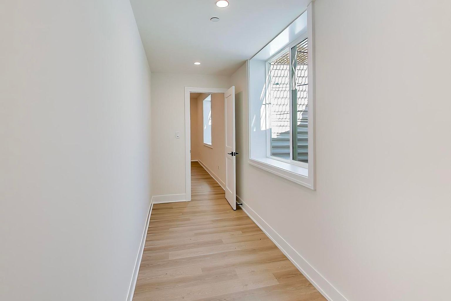 This is an interior shot of a bright hallway with light wood flooring and white walls. A window on the right side allows natural light to flood the space, and an open doorway leads to another room, creating a sense of depth. The hallway appears clean and modern, suggesting a well-maintained property.