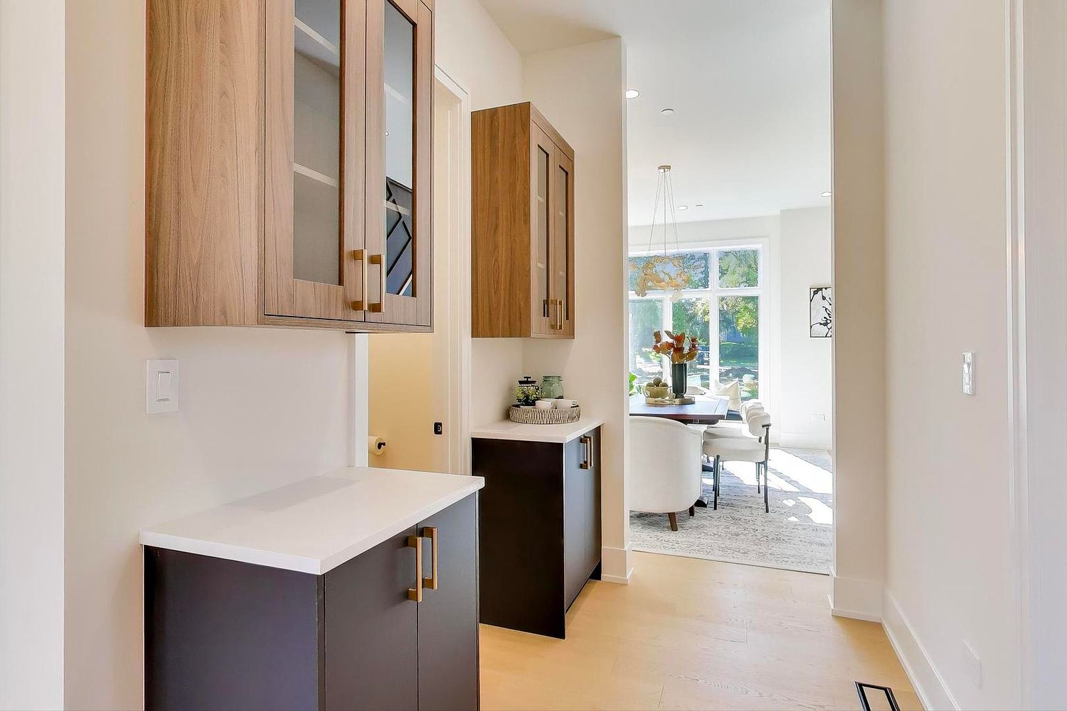 This interior shot showcases a hallway leading into a dining area. The hallway features custom cabinetry with contrasting wood and dark finishes, topped with white countertops. The dining area is bright and airy, with a large window providing natural light, and is furnished with a dining table and white chairs, creating an inviting and modern atmosphere.